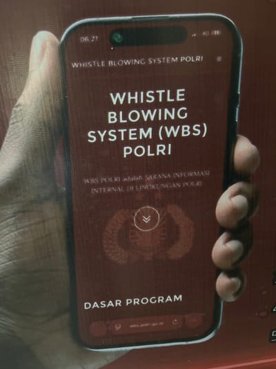 The image shows a close-up of a person's hand holding a smartphone. The phone displays a red screen with white text. The top of the screen shows the time "06.21" and a green dot indicating a camera. The text on the screen reads "WHISTLE BLOWING SYSTEM POLRI", and below that, "WHISTLE BLOWING SYSTEM (WBS) POLRI". Further down, there is smaller text that reads "WBS POLRI adalah SARANA INFORMASI INTERNAL DI LINGKUNGAN POLRI". At the bottom of the screen, the text "DASAR PROGRAM" is visible, along with a URL that appears to be "wbs.polri.go.id". The background is a blurry reddish-brown texture, possibly a surface or fabric. The location of Tanakawu, Indonesia, is not directly depicted or implied by the visual elements in the image.