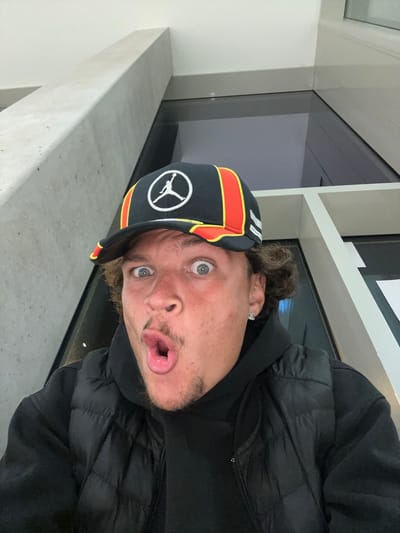 A selfie was taken indoors, apparently in the city of Ivry-sur-Seine, France. The subject is a man wearing a black, orange, and red hat with a white logo, a black hoodie, and a black puffer jacket. His face is contorted with wide eyes and an open mouth. The background includes a white shelf and a dark window. The camera angle is slightly tilted.