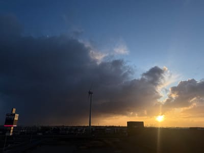 This image depicts a panoramic view of an industrial park in Naaldwijk, Netherlands, during sunset or late afternoon. The sky is characterized by a stark contrast: a substantial, dark grey cloud mass dominates the left half, appearing to bring heavy weather and subtly revealing a faint, circular object (likely the moon) through its density in the upper left. The right half of the sky exhibits scattered clouds illuminated by the low-lying sun, casting a strong golden-orange hue, with clear blue sky visible towards the top right.
A tall, three-bladed wind turbine, light grey in color, stands prominently slightly left of the image's center, its upper portion extending into the dark cloud mass. To the far left, a tall, multi-panel advertising signpost, partially shadowed but with illuminated sections, displays text including company names such as "SIZO," "TRADIRO," and "TanGO." Below the horizon line, the landscape is occupied by numerous low-rise industrial and commercial buildings, along with visible roads and infrastructure. The immediate foreground consists of a dark, elevated landmass. No individuals or active vehicles are discernible. The scene is observed from an elevated perspective, overlooking the `Bedrijvenpark Honderdland` industrial area, likely facing an easterly or southeasterly direction.