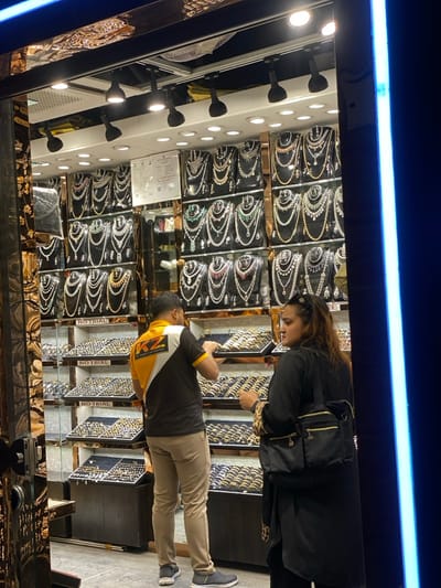 The image captures the interior of a jewelry store in Narayanganj, Bangladesh. A male individual, identified as an employee by his black and yellow "KZ JEWELLERS" polo shirt, stands with his back mostly to the camera, leaning over and interacting with items in a glass display case filled with rings or bangles. He wears light brown trousers and dark athletic shoes. Adjacent to him on the right, a female customer, dressed in a long black garment over a leopard-print top and carrying a black shoulder bag, stands facing slightly away from the male and towards the right of the frame.

The store's interior is brightly illuminated by overhead track lighting with multiple spotlights and recessed ceiling lights. The background features numerous wall-mounted display units showcasing a large collection of necklaces, many with white and colored stones, arranged on black busts. Below these, multiple tiered glass display cases are filled with rings, bangles, or similar small jewelry items. Some shelves within these cases bear "NO TRIAL" signs. A security camera (CCTV) is visible on the back wall, along with a white rectangular sign displaying text and a QR code. The left side of the frame shows a dark, reflective architectural element with ornate gold-colored patterns, likely part of the store entrance or facade. A vertical blue LED light strip runs along the far right edge of the image.