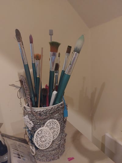 The image shows a close-up view of a woven cylindrical container filled with various art brushes. The brushes have different bristle types, sizes, and handle colors, with some appearing to be made of wood and metal. The container itself is wrapped in a textured yarn in shades of grey, white, and brown. Several decorative items are attached to the outside of the container, including white circular lace-like ornaments and a single pearl on a dangling chain. A blue USB cable is also plugged into the container's rim. The background is a plain, light-colored wall with subtle shadows cast upon it. The scene is set on a light-colored surface, possibly a shelf or table, with a small pink object visible in the lower right corner. The image does not provide any information about the location in the city of Waverley, United Kingdom.