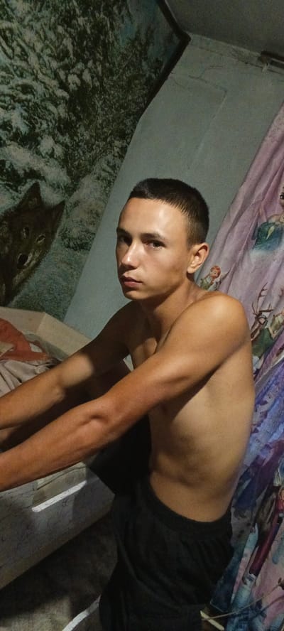A young man, appearing to be in his late teens or early twenties, is seated indoors and looking towards the camera. He is shirtless and wearing dark shorts. His hair is short and neatly cut. The background shows a wall with a mural or tapestry of a wolf on the left and what appears to be a shower curtain or fabric with cartoon characters on the right. The lighting suggests it is daytime, though the indoor setting makes it difficult to ascertain the exact time of day. The overall mood is relaxed and casual. There is no visible text in the image.