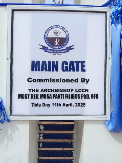 The image shows a white plaque mounted on a white wall with blue ribbon decorations on either side. The plaque is framed in silver and reads "MAIN GATE Commissioned By THE ARCHBISHOP LCCN MOST REV. MUSA PANTI FILIBUS PHD. OFR This Day 11th April, 2025". The top left corner of the plaque features a circular emblem with the words "BRANHUM LUTHERAN SEMINARY" written around its circumference. The emblem also includes a cross symbol, the letters "LCCN" and "VOLA". The inscription below the emblem reads "TO SERVE GOD AND HUMANITY EST. 1985". The coordinates 9.187, 12.525 correspond to the location of Rumde Waziri in Nigeria. The plaque is likely commemorating the opening of the main gate to the Branhum Lutheran Seminary.