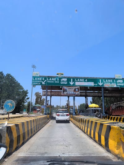 The image shows a toll plaza on a clear day. The toll plaza has multiple lanes, indicated by signs labeled "LANE 6," "LANE 5," and "LANE 3." Lane 6 is designated for "OVERSIZE VEHICLE," while lanes 5 and 3 are for "TRUCK/BUS." Some lanes also indicate "NO CASH ONLY & UPI" for payment methods. A white SUV is visible in one of the lanes, and other vehicles, including a truck, are also present. In the foreground, there are concrete barriers painted with yellow and black stripes. A blue circular sign with an image of a helmet and the text "YOU MUST WEAR YOUR HELMET" is visible on the left side of the road. The background shows trees and a clear blue sky. The location is identified as Chersoo, India.