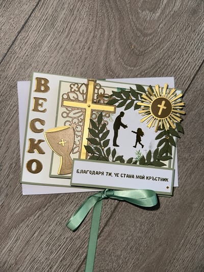 This is a close-up, high-angle shot of a handmade card on a wooden surface. The card is decorated with gold-colored embellishments and text. On the left side of the card, there are five large gold letters spelling "ВЕСКО" vertically. To the right of these letters, there is a gold chalice with a cross inside it. The central part of the card features a large gold cross with intricate cut-out details. Surrounding the cross are dark green paper leaves, some adorned with tiny gold dots. On the right side of the card, there is a gold sunburst emblem with a cross in the center. Below this, a silhouette of a man reaching out to a child is depicted. A white rectangular banner with gold Cyrillic text that reads "БЛАГОДАРЯ ТИ, ЧЕ СТАНА МОЙ КРЪСТНИК" is placed across the bottom of the card. A mint green ribbon is tied in a bow at the bottom of the card, with its tails extending downwards. The wooden surface appears to be a floor or tabletop with visible grain patterns. The lighting is even, and the focus is sharp on the card. There is no identifiable location in Belfast City, United Kingdom.