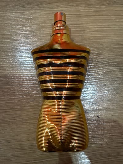 A gold and brown striped perfume bottle with a gold cap is positioned vertically on a wooden surface. The bottle has a torso-like shape, with horizontal stripes of gold and dark brown. The wooden surface has a visible grain pattern and reflects light. There is no indication of the city of Dolna Banya, Bulgaria, or any people in the image.