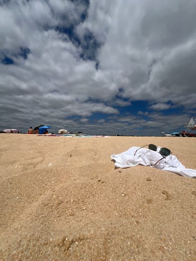This image captures a beach scene in Albufeira, Portugal. The perspective is low, taken from the sandy ground. In the immediate foreground, a crumpled white fabric item, possibly a shirt, lies on the light brown sand, with a pair of sunglasses resting atop it. The sunglasses feature dark, reflective lenses and visible dark frames with red and green striped temples.

The midground expands into a wider stretch of sand where several groups of individuals are present. Approximately five to ten people are visible, some sitting or reclining under beach umbrellas. A distinct blue beach umbrella is centered, with individuals gathered beneath it. To the left, another person is partially visible in red swimwear near a light-colored canopy. Towards the far right midground, a black and white striped beach umbrella is erected. Various beach towels, mats, and other personal belongings are scattered on the sand around these groups.

The horizon line is relatively flat, and the distant background appears indistinct, possibly showing the far end of the beach or distant coastal features. The sky above is characterized by a significant presence of grey and white cumulus or stratocumulus clouds, interspersed with patches of clear blue sky. The overall lighting suggests daylight conditions. No distinct actions or interactions beyond general beach relaxation are clearly discernible among the distant figures.