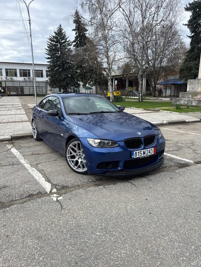The scene is located in Bregovo, Bulgaria, depicting an outdoor parking area on an overcast day. A dark blue two-door BMW E92 coupe is parked at an angle in the foreground, facing slightly left. The vehicle features a black grille, aftermarket silver multi-spoke BBS wheels with visible red brake calipers, and a black front lip spoiler. Its front license plate displays "BG 815 M 243" within a custom "Born in Town" frame, with "26" visible in the bottom right corner of the plate. The parking surface is asphalt, exhibiting cracks and faded white parking space markings.
In the background to the left, a two-story white building with multiple windows, some fitted with vertical bars, is visible. Several air conditioning units are mounted on its lower facade, which features a grid or patterned paneling. A paved walkway runs in front of this building. A tall metal street light with draped string lights stands in the left midground. Two tall, dark green coniferous trees are positioned behind the street light.
To the right of the vehicle, a park-like area contains several bare deciduous trees, indicative of late winter or early spring. A children's playground with a yellow slide and swings is partially visible. Further right, a concrete structure, possibly a monument base, is present. Behind the playground, a derelict or unfinished building with exposed concrete pillars, brickwork, and a blue tarp covering part of its structure can be seen. No individuals are present in the scene.