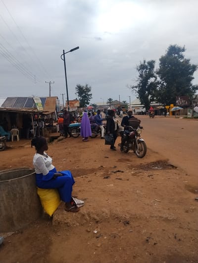 In Mangu Town, Nigeria, a woman sits on a yellow sack next to a concrete structure, wearing a white top and blue trousers. The scene is an outdoor market with several people and motorcycles. In the background, there are shops with solar panels on their roofs, a lamppost, and trees under an overcast sky. A motorcycle with a rider and passenger is parked on the dirt road. Other individuals are seen walking or standing around.