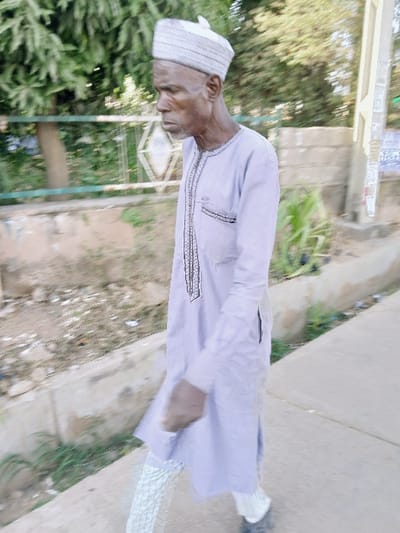An elderly man with a traditional cap and light-colored robe stands outdoors. His skin is dark, and his features are aged. He is next to a sidewalk and some greenery. A white structure with a decorative design is visible behind him, along with trees and plants. The background is slightly blurred, giving prominence to the man in the foreground.