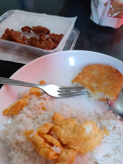 The image shows a plate of food on a dark surface. The plate is white and contains a portion of white rice, scrambled eggs, and a fried potato patty. A silver fork is lying on the plate near the edge of the potato patty. A clear plastic container with leftover fried chicken is in the background. This location in Parañaque, Philippines, is a residential area with small homes and businesses.