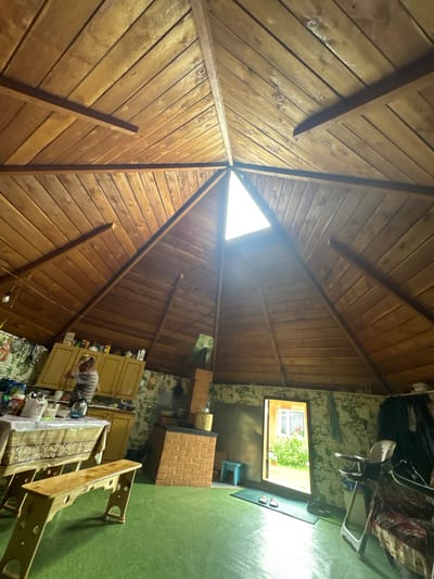 The image shows the interior of a rustic, wooden structure with a conical roof supported by beams. Natural light enters through a triangular opening at the apex of the roof. The walls are covered with a green, possibly painted, textured material, with visible brickwork in places, particularly around a large brick oven or fireplace. In the foreground, a light brown wooden bench with circular cutouts sits on a green floor. To the left, a table is covered with a patterned tablecloth, with dishes and containers on it, and a person wearing a striped shirt is visible in the background near kitchen cabinets. Further to the right, a doorway opens to an exterior scene of lush greenery and a building with windows. A baby high chair is positioned to the right of the doorway, and other items, including dark fabric and a chair with a patterned cover, are visible in the same area. The scene appears to be a dwelling or an outdoor covered space, possibly a kitchen or a dining area. The location is identified as Yabogan, Russia.