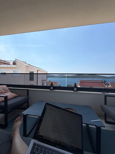 A first-person perspective shot captures a person's bare foot, with dark nail polish, resting on a grey sofa on a balcony. The person is working on a silver laptop, open and positioned on a dark grey coffee table. The laptop screen displays text in a foreign language. The balcony offers a view of the city of Šibenik, Croatia, with buildings featuring red-tiled roofs. Beyond the city, a body of water is visible, with a distant island on the horizon under a clear blue sky. The scene appears to be during daytime.