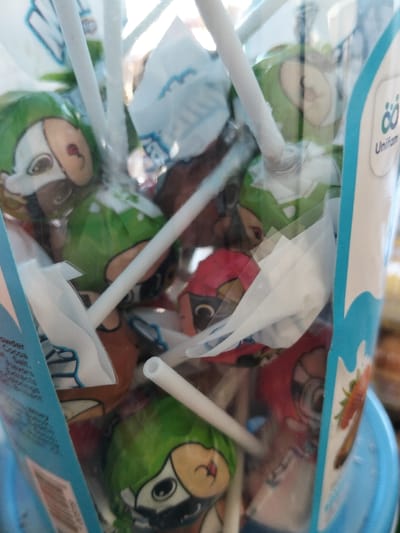 The image displays a close-up view of numerous lollipops contained within a clear plastic dispenser or jar. The lollipops are individually wrapped in colorful plastic, featuring cartoon characters. Many of the wrappers are green, depicting a character that appears to be a dog or animal with large, dark eyes, a small brown nose, and a white face covering or snout area, wearing a green hooded outfit. Other lollipops are wrapped in red and brown, showing characters of a similar cartoon style. Each lollipop has a white, presumably plastic, stick, some of which appear to be hollow.
The container itself has blue branding elements on its sides. On the right, a white logo resembling two interconnected loops or an infinity symbol is visible above the word "Unifam." Further down on this side, a graphic of a red strawberry is present, next to the word "Strawberry." On the left side of the container, a partial ingredients or nutritional label is visible in white text against the blue background. Words that can be discerned include "Powder," "Cocoa," "Salt," "Flavors," and "Contains." The lollipops are densely packed, filling the container, with many sticks pointing in various directions. The overall lighting is bright, causing some glare and reflections on the plastic container and wrappers. The background behind the container is out of focus, suggesting the lollipops are the primary subject of the photograph.