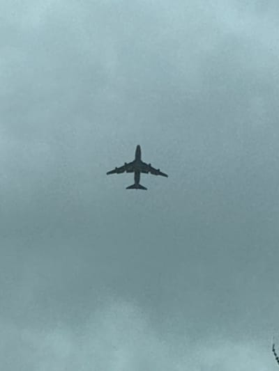 The image depicts a solitary four-engine, wide-body aircraft, rendered as a dark silhouette, centered against a uniformly light grey, overcast sky. The aircraft is viewed from its ventral aspect, showcasing its wings, fuselage, and four distinct engine nacelles. No individuals or ground-based structures are visible. The scene is observed over Berg, Luxembourg.