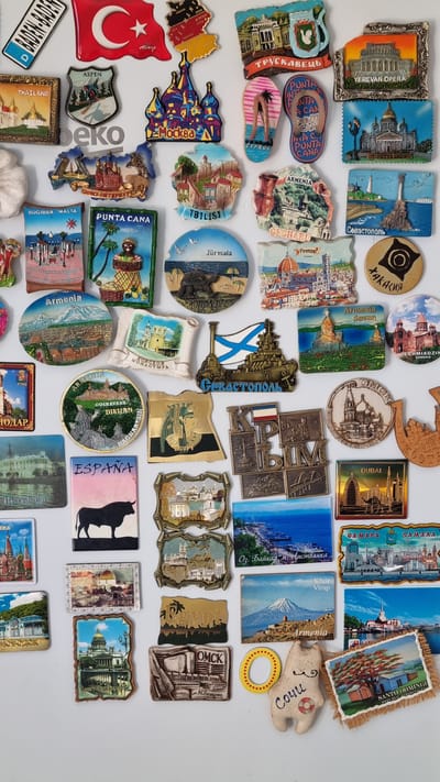 The image is a close-up, flat lay photograph of a collection of refrigerator magnets displayed on a white surface. The magnets depict various landmarks and scenes from different locations, primarily tourist destinations. Many of the magnets feature text in Cyrillic and other languages, indicating their origins. Some of the visible locations include Turkey (flag), Germany (map), Moscow (St. Basil's Cathedral), Thailand, Cyprus (flip-flops), Saint Petersburg (Winter Palace), Sevastopol, Egypt, Armenia, Dubai, and Samara. There are also magnets with general scenes like mountains, beaches, and cityscapes. The magnets are varied in shape, size, and material, with some appearing to be made of metal and others of plastic or resin. In the lower right quadrant, there is a small fabric plush toy resembling a cat with the word "Сочи" (Sochi) embroidered on it. The image does not contain any people, actions, or interactions, and the environment is a plain white background. There is no depiction of the city of Chereshnya, Russia.