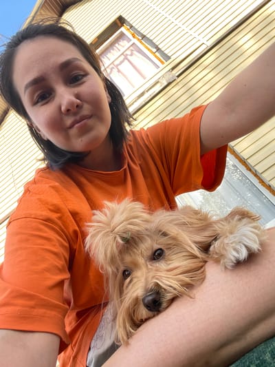 A woman in an orange t-shirt is holding a small, tan-colored dog. The dog is resting on the woman's leg, looking at the camera. The background consists of a window with visible shutters. The location is implied to be Verkh-Katunskoye, Russia.