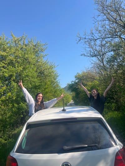 Two women stand beside a white car on a grassy area with trees and a clear blue sky above. Both women extend their arms and make peace signs with their fingers. The location is Rish, Bulgaria, as inferred from the image context.