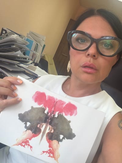 A woman wearing large, black-rimmed glasses, a white top, and large earrings holds a Rorschach inkblot test card in her right hand. Her nails are manicured with a gradient design. She is surrounded by a cluttered stack of papers and documents. She has a tattoo on her upper arm. The setting is inside a room with light yellow walls, and the presence of files suggests an office or study environment. There is no evidence to suggest the location is Murmansk, Russia.