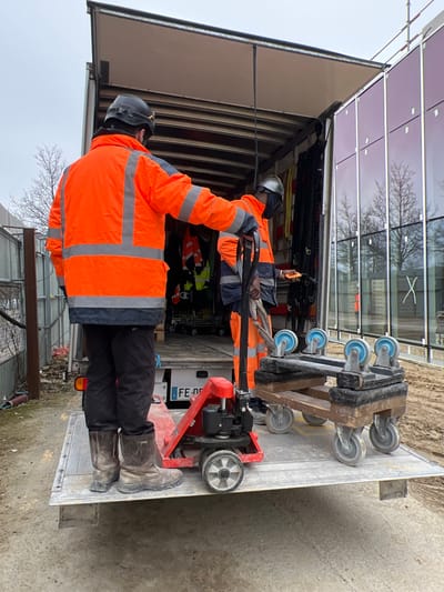 This image depicts a construction or industrial site in Aulnay-sous-Bois, France. Two male workers are engaged with the rear of a white box truck.

The worker in the foreground stands on the truck's lowered liftgate, with his back to the camera. He wears a dark hard hat with a yellow accent, a bright orange high-visibility jacket with grey reflective strips, dark trousers, and dark brown work boots. He is holding the handle of a red manual pallet jack, which is positioned on the liftgate.

The second worker is partially visible inside the truck's cargo area, facing towards the right. He wears a protective face shield, an orange high-visibility garment with blue/grey accents, and dark trousers. He holds a small orange device, possibly a remote control or scanner, in his right hand and a bundle of heavy cables or straps in his left hand. The truck's interior contains various pieces of equipment, including hanging high-visibility clothing. The French license plate "FE-007..." is partially visible on the truck's rear.

To the right of the pallet jack, on the liftgate, are two multi-wheeled industrial dollies or carts, stacked or placed side-by-side, featuring multiple blue casters.

On the far right, a modern building with large, reflective glass panels, some with a purple/red tint, is visible. The glass reflects bare trees and an overcast sky. A white "X" is marked on one of the glass panels. Scaffolding or structural elements are present above the building. The ground surrounding the truck is unpaved, appearing as dirt or compacted soil. A metal fence or barrier is visible on the far left of the frame. The sky is overcast.