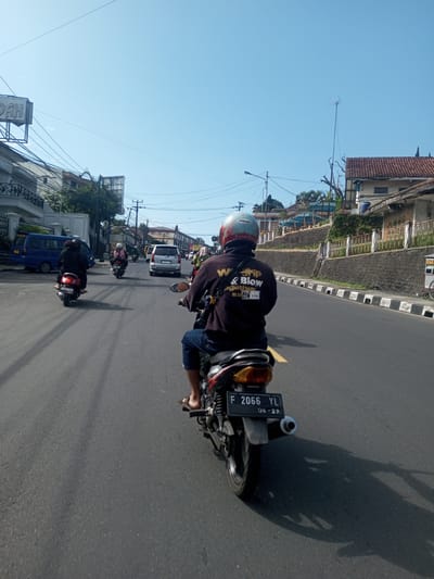 The image, taken from the perspective of a person riding a motorcycle, shows a street scene with other motorcyclists and vehicles on a paved road under a clear blue sky. The rider in the foreground is wearing a dark long-sleeved shirt with text on the back that includes the words "Wdrip & Blow together" and a helmet. The motorcycle has a license plate reading "F 2066 YL 06-27". The environment consists of buildings, utility poles, and other street elements suggesting a moderately populated area. Based on the OCR text, this image appears to be taken in Indonesia, as the license plate format is commonly associated with Indonesian vehicle registration. Laos has not been identified in this image.