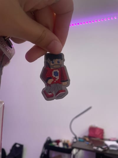 A person's hand holds a small, blocky figurine with a red jacket and gray pants. The figurine resembles a character from a video game. In the background, a string of pink LED lights runs horizontally along the wall. The scene is indoors, and the lighting suggests it is nighttime or a dimly lit room. The location of Лесное Снт, Russia is not depicted or inferable from the image.