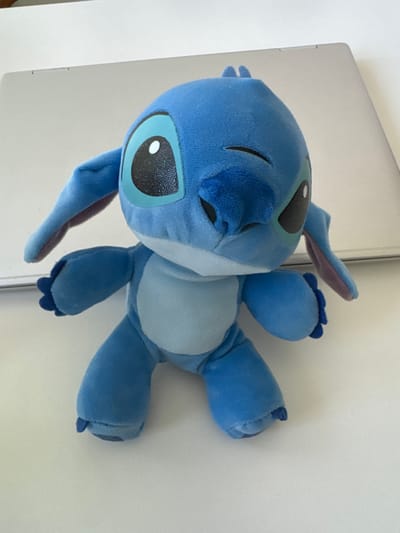 A stuffed toy resembling the character Stitch from Disney's "Lilo & Stitch" sits on a white surface, with the edge of a silver laptop visible in the background. The toy is predominantly blue, with large black eyes that have a light blue outline and a white pupil. One eye is closed in a wink. Its ears are long and floppy, with a lighter purple-ish underside. The plush toy is in a standing pose, with its arms slightly extended and legs bent. The surface it rests on is white and appears to be a table or desk. The environment suggests an indoor setting, likely a home or office, with no specific landmarks visible that would indicate a location in Winterthur, Switzerland. The lighting is bright, casting subtle shadows on the surface around the toy.