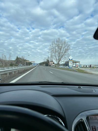 The image provides a first-person perspective from the interior of a vehicle, looking forward through the windshield onto a multi-lane highway in Tserovo, Bulgaria. The vehicle's black dashboard and a portion of the steering wheel are visible in the foreground. A digital display on the dashboard shows "08:32".
The highway consists of at least two lanes in the direction of travel, marked by white lines, and is bordered by a metal guardrail on the left. The sky is overcast with a dense pattern of altocumulus clouds. The surrounding vegetation, including a large, bare deciduous tree on the right side of the road, indicates a non-growing season.
On the right side of the highway, a Lukoil gas station with a distinctive blue and yellow canopy and signage is clearly visible. Several passenger vehicles are parked near the gas station pumps. Beyond the gas station, a line of heavy goods vehicles, some marked with "GÖKSEL," are parked at what appears to be a truck stop or rest area. A red and white circular road sign is also visible near the gas station entrance.
On the left side of the highway, beyond the guardrail, sparse bare trees and brush line the road. Distant vehicles are visible traveling on the highway in both directions. A large gantry structure spans the highway further in the distance. No specific individuals or active interactions are clearly discernible.