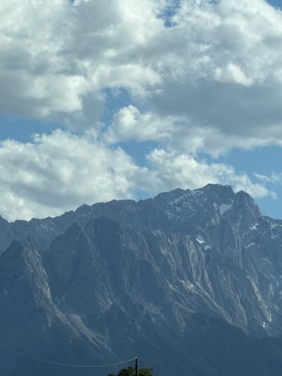 The image depicts a view of the Zugspitze, Germany's highest peak, as seen from Farchant, Germany. The mountain range dominates the lower half of the frame, with jagged, gray rock formations interspersed with patches of snow. A structure, likely a weather station or observation tower, is visible on the summit. The sky above is a mix of blue and white clouds, with some of the clouds appearing heavy and textured. A thin wire stretches horizontally across the lower portion of the image, and a dark green tree is partially visible at the bottom. The overall impression is of a majestic mountain landscape under a cloudy sky.