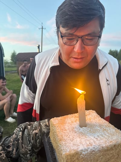 An outdoor, close-up shot shows a man in his 30s with dark hair and glasses blowing out a single lit candle on a rectangular cake. He is wearing a black t-shirt and a white and black jacket with red accents. The cake is dusted with powdered sugar. In the background, two other shirtless men are visible near a wooden structure, possibly a sauna, and some trees. The setting appears to be a rural or semi-rural area, consistent with the outskirts of Yakty-Kul, Russia, given the presence of natural elements and simple structures. The sky is overcast with a hint of sunset colors. The focus is on the man and the cake, with the background blurred.