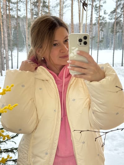 An adult female is depicted taking a selfie indoors, looking slightly to her right towards the smartphone held in her right hand. The phone has a light-colored, possibly glittery, case and a triple-lens camera system. Her left hand is near the hood of her light cream-colored puffy jacket, which is worn over a pink hooded sweatshirt. The jacket features small, light blue embroidered dots. She has light brown hair, small earrings, and light pink fingernails. The background, viewed through a large window or glass pane, reveals a snowy winter forest scene in Mishkino, Russia, with numerous bare deciduous trees and evergreen pines, and snow covering the ground under an overcast sky. In the foreground, a branch with yellow, fluffy blossoms is visible on the left, and a dark, bare branch extends into the frame on the right.