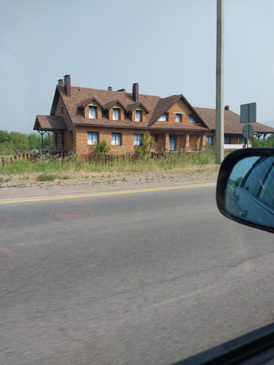 The image shows a multi-story brick building with a brown shingle roof, viewed from the side of a road. The building has many windows and several dormer windows. There is a covered porch area on the ground floor. Greenery, including trees and bushes, surrounds the building. The road in the foreground is asphalt with a yellow line painted down the center. Red markings are visible on the road surface. The photo appears to be taken from a moving vehicle, as indicated by the reflection in the side mirror, which shows trees and sky. The sky is clear and light blue, suggesting it is daytime, likely with good weather. The overall scene is that of a building located in a rural or semi-rural area, possibly a hotel or resort given the architecture and setting, consistent with a location like Belokurikha, Russia. No people are visible in the image. There is no discernible text in the image.