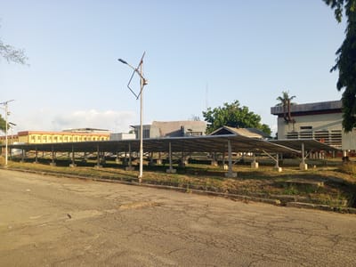 The image depicts a street scene in Tawpo, Nigeria, featuring a row of solar panels erected on concrete supports. The panels are situated on a grassy area adjacent to the road. Behind the panels are several buildings of varying architectural styles, including one with a distinct yellow facade. A street lamp stands near the panels, its design appearing incomplete. The road surface is cracked and shows signs of wear. The sky is clear and light blue, suggesting a daytime scene. Vegetation, including trees and grasses, are visible throughout the environment.