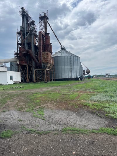 The image depicts an industrial scene in the city of Progress, Kazakhstan. In the foreground, a dirt and gravel area with patches of dry, cracked mud and sparse green grass is visible. A small, white building with a single window and a wooden bench sits to the left. Adjacent to the building is a large, weathered industrial structure with multiple towers, rusty pipes, and metal platforms, likely part of a grain processing or storage facility. To the right of this structure is a large, silver, corrugated grain silo. Pigeons are scattered across the grassy area in front of the industrial complex. In the background, more industrial buildings, a railway line, and distant structures are visible under a cloudy sky.