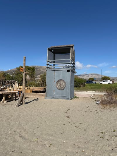 The image displays a tall, grey, box-like structure with an open, framed upper section, resembling a kiosk or small stand, positioned outdoors under a clear blue sky. A prominent circular, woven, target-like decoration is affixed to the side of the main body of the structure.

To the left of this structure, a wooden post stands, bearing a light-colored rectangular sign with the Greek word "Dromos" visible, which translates to "Road" or "Street."

The foreground consists of a wide area of sandy or gravelly ground. In the mid-ground, behind the grey structure, there are low bushes and some trees. Further back, on what appears to be a road or track, a white car and a dark-colored car can be seen. Rolling mountains dominate the distant background.

The scene is set on a bright, sunny day, likely midday or early afternoon, indicated by the clear sky with minimal clouds and strong illumination. The overall environment suggests a dry, outdoor setting, possibly in the Anatoliki Attiki region of Greece. There are no people visible, and no specific activities are taking place.