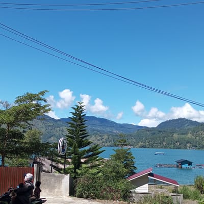 In the city of Kalalengkio, Indonesia, a vibrant scene unfolds under a clear blue sky with scattered white clouds. The foreground is dominated by a paved road where a person on a motorcycle, wearing a white helmet with a red and black design, is accompanied by a child. To the left, a red metal fence runs horizontally.
Further into the scene, a tall evergreen tree stands prominently, its branches reaching towards the sky. Behind it, a large body of water, likely a lake, shimmers with a bright turquoise hue. In the distance, rolling hills covered in dense green forests form a majestic backdrop, with layers of mountains receding into the haze.
On the water, several structures are visible. A small blue boat navigates the calm surface. Closer to shore, a blue-roofed floating structure with several compartments is anchored. Another, smaller floating structure with multiple colored buoys is also present.
To the right, a white building with a red roof is partially visible, suggesting a dwelling or a small establishment. The overall atmosphere is one of natural beauty and tranquility, with a touch of human presence.