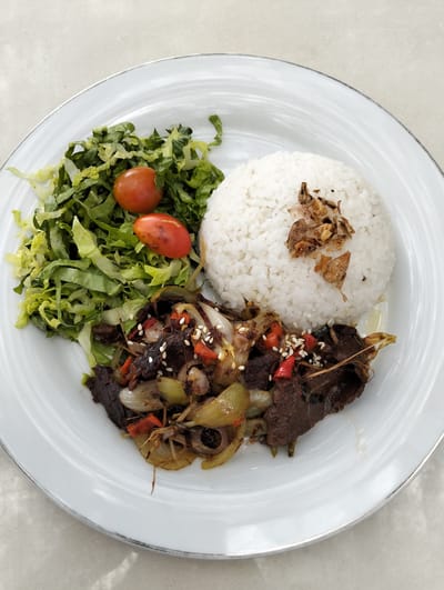 A white plate contains a meal on a textured, light-colored surface. The plate is divided into three sections. The top left section holds a pile of shredded green lettuce with two small, red cherry tomatoes on top. The top right section features a mound of white rice, topped with fried shallots. The bottom section contains a stir-fry of dark-colored meat, sliced onions, and red chili peppers, garnished with sesame seeds. The overall scene is a close-up, overhead shot of food, suggesting it was taken in a restaurant or home setting. The lighting appears to be natural, possibly from a window, casting subtle shadows. There are no people or animals visible in the image. The image appears to be a product photo for a dish from a restaurant in Cikole, Indonesia, as suggested by the context.