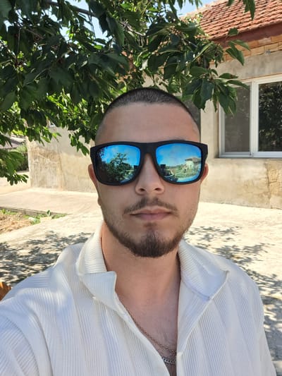 A medium shot captures a young man with a short buzz cut and a beard, wearing black sunglasses and a white textured shirt with a gold chain. The sunglasses reflect a rural scene with a house and trees. He is outdoors, with dappled sunlight creating shadows on the ground. In the background, a pale yellow building with a red-tiled roof and a window is visible, partially obscured by the branches of a leafy green tree. The location appears to be Ignatievo, Bulgaria, based on the architecture and vegetation.