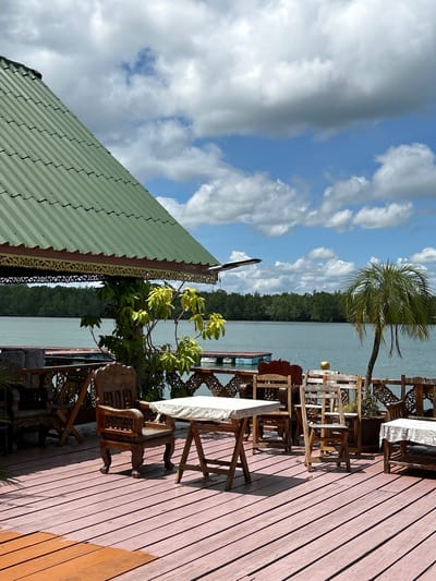 This image depicts an outdoor seating area at a restaurant located on a pier in Tambon Ko Panyi, Thailand. The area is furnished with several wooden tables and chairs, some covered with white tablecloths. A green-tiled roof with ornate trim extends over part of the seating area, providing shade. In the background, a body of water is visible, bordered by a dense, green treeline. The sky is partly cloudy. A single palm tree stands to the right of the seating area. The overall impression is a casual, waterside dining setting.