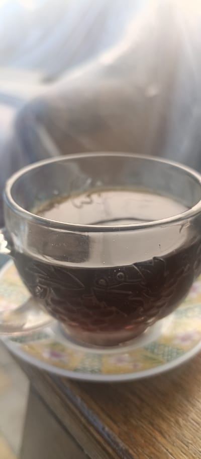 A close-up, eye-level shot shows a clear glass teacup filled with a dark liquid, likely tea, sitting on a saucer. The teacup has a decorative, embossed pattern that appears to depict dragons or birds. Faint steam rises from the surface of the liquid. The saucer has a patterned rim with yellow and green diamond shapes and small floral accents in pink and purple. The teacup and saucer are placed on a wooden surface, possibly a table. The background is softly blurred, suggesting an indoor setting with diffused light, possibly near a window. The scene does not provide any specific details about the city of Voskresenskiy, Russia, or any people.