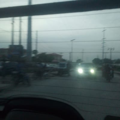 The image, likely captured from inside a vehicle, depicts a street scene in Isunba, Nigeria. The visibility is obscured, suggesting possible weather conditions like fog or haze. Traffic, including a car with headlights on and a motorcycle, is visible on the road. Overhead power lines and utility poles are present. Buildings and trees are discernible in the background, albeit blurred. The foreground consists of interior elements of the vehicle, such as the dashboard.