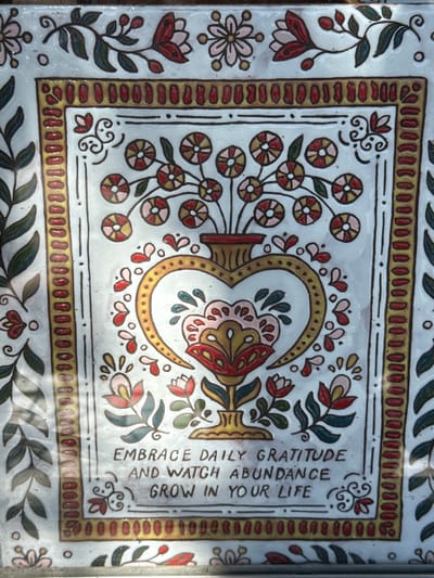 The image shows a painted tile, possibly ceramic, featuring a symmetrical floral design. The design includes stylized flowers, leaves, and a central heart-shaped element, all rendered in red, gold, green, and pink hues. The edges of the tile are bordered with a repeating pattern of oblong shapes. On the right side of the tile are inscribed the words, "EMBRACE DAILY GRATITUDE AND WATCH ABUNDANCE GROW IN YOUR LIFE." This style of folk art is prevalent in Salta, Argentina, though specific location is unknown from the image. The tile has an overall aesthetic of handmade craftsmanship with slight imperfections in the paint application. It appears to be mounted as part of a larger surface adorned with similar tiles and designs.