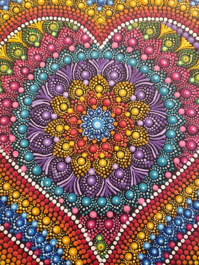 A close-up view of a vibrant, colorful dot mandala. Concentric circles of blue, orange, yellow, purple, pink, and green dots radiate from the center. Intricate patterns and radial symmetry are created with the various colored dots. Black background separates the patterns. The use of white dots creates the effect of diamonds and further emphasizes the layers and symmetry of the design.
