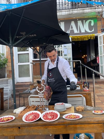 A man, wearing a black apron with the logo "Jamónpassion EXCLUSIVE EVENTS", is slicing a large cut of ham with a knife and fork. He is standing behind a wooden table laden with plates of thinly sliced ham and breadsticks. The scene appears to be outdoors, possibly at a restaurant or event. A large black umbrella, with the "FEVER TREE MIX WITH THE BEST" logo, provides shade. In the background, a sign reads "BLANC" in large white letters. The floor is paved with reddish-brown tiles. The overall setting suggests a food-related event or establishment in Arenys de Mar, Spain.
