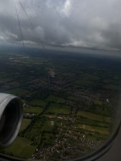 A view from an airplane window shows green fields and the edge of a town, likely in the Reigate and Banstead area of the United Kingdom. The sky above is overcast with gray clouds. Part of the airplane engine is visible in the lower right corner. The plane appears to be landing or taking off, providing a clear view of the landscape below.
