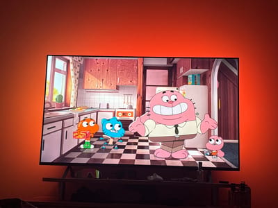 The scene depicts a large flat-screen television displaying an animated cartoon, set within a domestic environment in the city of Straldzha, Bulgaria. The television is mounted on a dark media console, and the wall behind it is bathed in a vibrant orange-red ambient light.
On the television screen, an animated kitchen is depicted with a black and white checkered floor, white tiled countertops and backsplash, light wooden upper cabinets, and a window on the left revealing greenery outside and a potted cactus on the sill. Four anthropomorphic characters are present. A large, pink rabbit wearing a tan shirt and brown trousers stands in the foreground, smiling widely with open hands. To its left, a smaller blue cat stands in a crouched pose, and further left, an orange goldfish wearing green boots holds a yellow box labeled "#HA BROWNIEZ". On the far right, a very small pink rabbit, wearing an orange dress, holds a small white remote or device. A white refrigerator and a freestanding oven are also visible in the background of the cartoon scene.
Below the television, on the dark media console, a pair of white athletic shoes are positioned on the right. To the far right, several clear glass bottles or dispensers are arranged. A dark, indistinct object, possibly fabric, rests on the left side of the console. The television's stand is visible beneath the screen. The wall is otherwise unadorned, reflecting the intense orange-red illumination from behind the television. A dark, blurred object is partially visible in the bottom-left foreground, out of focus.