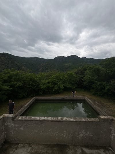 The image depicts an outdoor scene with a rectangular concrete basin filled with green water in the foreground. Two individuals are visible. To the left, a man stands near the edge of the basin, looking towards the camera. He is wearing a dark t-shirt with white stripes on the sleeves and dark pants. To the right, another person, possibly a woman, is standing on the edge of the basin, with their back to the camera, in a jumping or dancing pose.

The basin is situated on a grassy area, with a dense line of green trees forming a backdrop. Beyond the trees, a mountainous landscape with rocky peaks under a cloudy, overcast sky dominates the background. The overall atmosphere is natural and slightly subdued due to the cloudy weather. The image appears to have been taken in Akhtala, Armenia, given the context of the location.