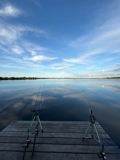 The image depicts a dock extending into a body of water, possibly a lake or river, under a blue sky with scattered clouds. Two fishing rods are positioned on the dock, set up with fishing line extending into the water. The water reflects the sky and the distant horizon, creating a mirrored effect. The dock is made of wooden planks. The location could potentially be Khryashchevka, Russia, based on user information. No people are visible in the image.