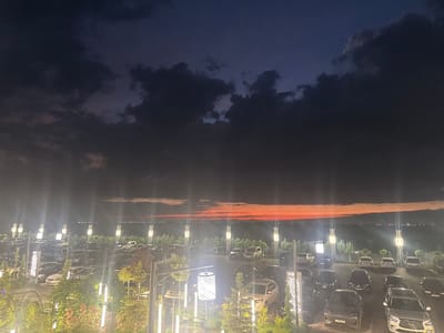 The image depicts a twilight scene in a parking lot with a dramatic sky. The sky is a deep, moody blue with dark, heavy clouds, but a vibrant band of orange and red hues stretches across the horizon, suggesting a sunset or the lingering light of dusk. The parking lot is filled with numerous cars, predominantly SUVs and sedans, suggesting a gathering or an event taking place at the location. Many of the cars are modern and well-maintained.

Several tall, illuminated poles with decorative lamps are spaced throughout the parking lot, casting a warm glow and highlighting the vehicles and surrounding trees. In the foreground, there are also smaller, modern outdoor light fixtures and some greenery, including trees and shrubs, some of which appear to be illuminated. The overall atmosphere is a mix of anticipation and the quiet beauty of twilight.

The scene is set in a location that appears to be an entertainment venue or a restaurant with ample parking, given the illuminated features and the density of cars. The vast, open landscape visible in the distance, under the colorful sky, hints at a location on the outskirts of a town or city. The time of day is clearly evening, transitioning from sunset into night.

One notable detail is a vertical sign with the word "VIP" in white, illuminated lettering, indicating a designated area or special access within the parking lot. There is also visible text on the license plates of some vehicles, though it is too small to be legible. The lighting and the colorful sky evoke a sense of calm and perhaps a touch of elegance.