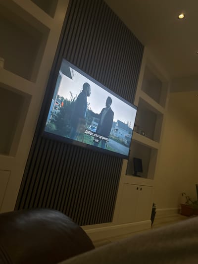 The image shows a television mounted on a wall with horizontal striped paneling. The TV screen displays a scene with two figures in a green, outdoor setting. Subtitles in a language other than English are visible. The wall features white, built-in shelving and cabinets. A chair is partially visible in the foreground. The image does not provide enough information to determine the location in the city of Chatham, United Kingdom.